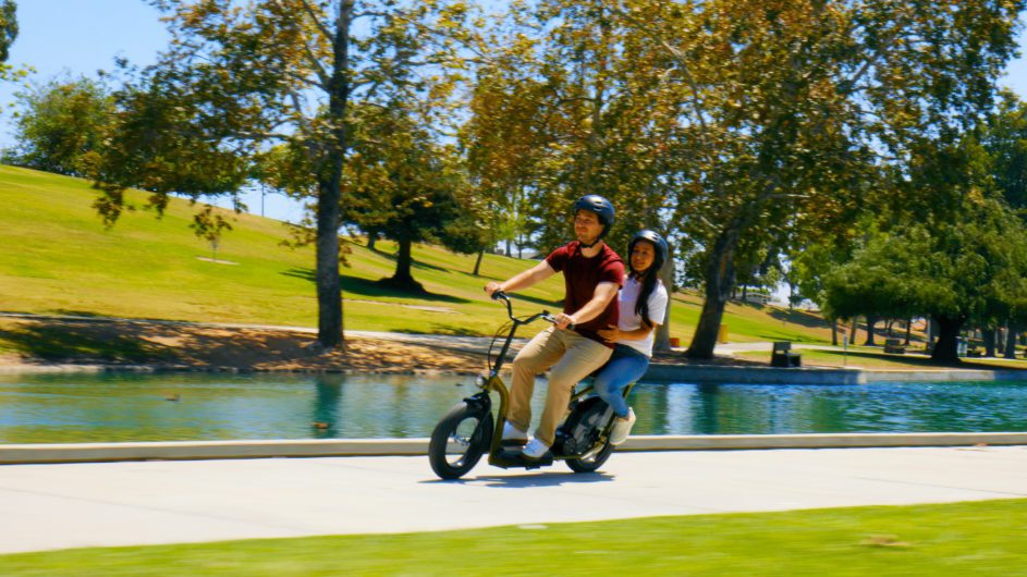 Two people riding a razor ecosmart scooter by a pond with trees in the background
